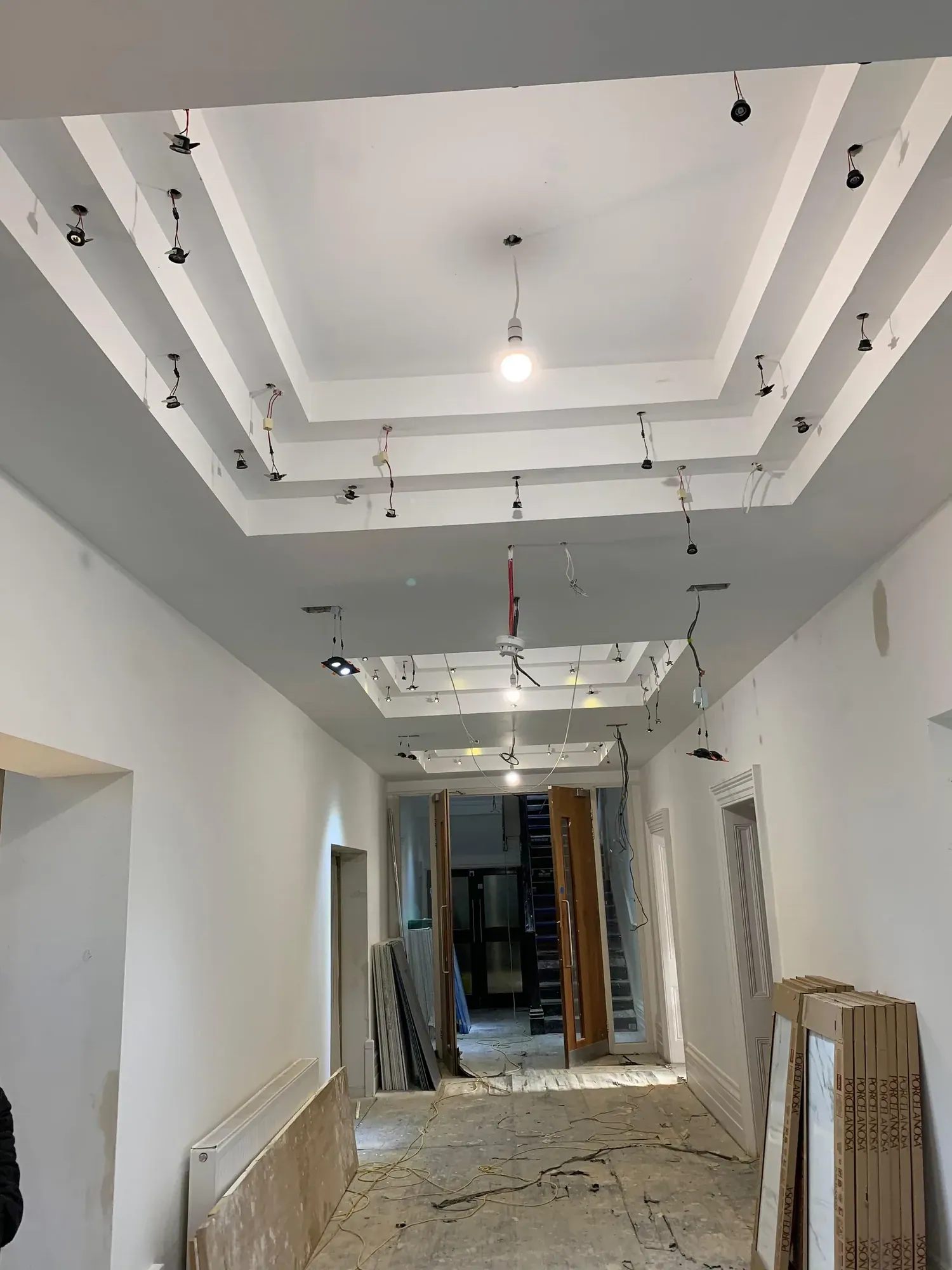 Corridor with a stepped plasterboard ceiling raft and recessed perimeter detail, pendant cable hanging, boards stacked at the end