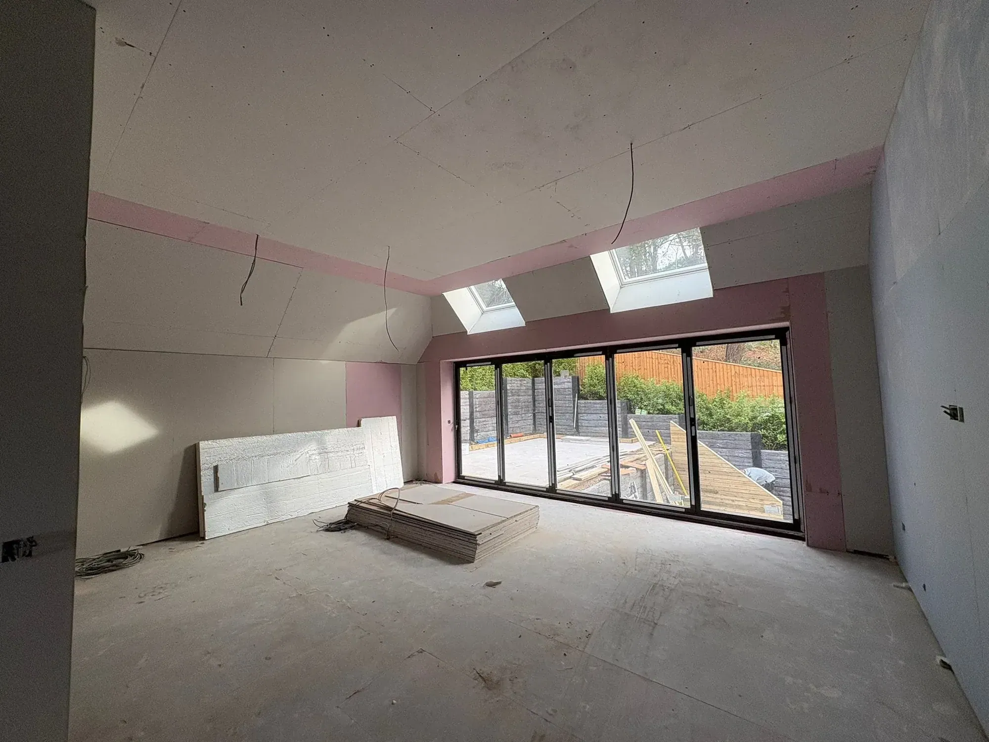 Single-storey extension boarded out, two rooflights in the sloping ceiling, four-panel bifold doors to the garden, stacked plasterboard on the floor