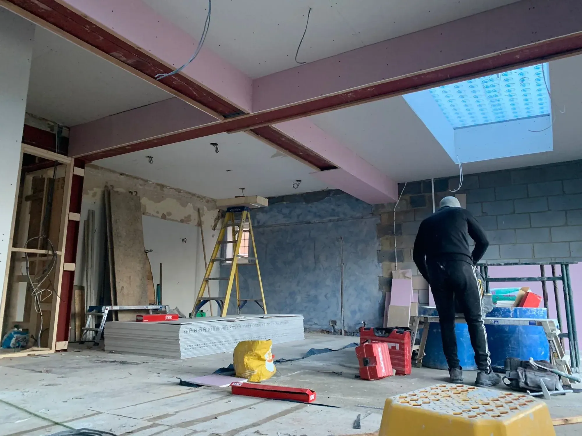 Operative on site beneath pink-painted steel beams and a rooflight, plasterboard stacked ready, blockwork wall still exposed