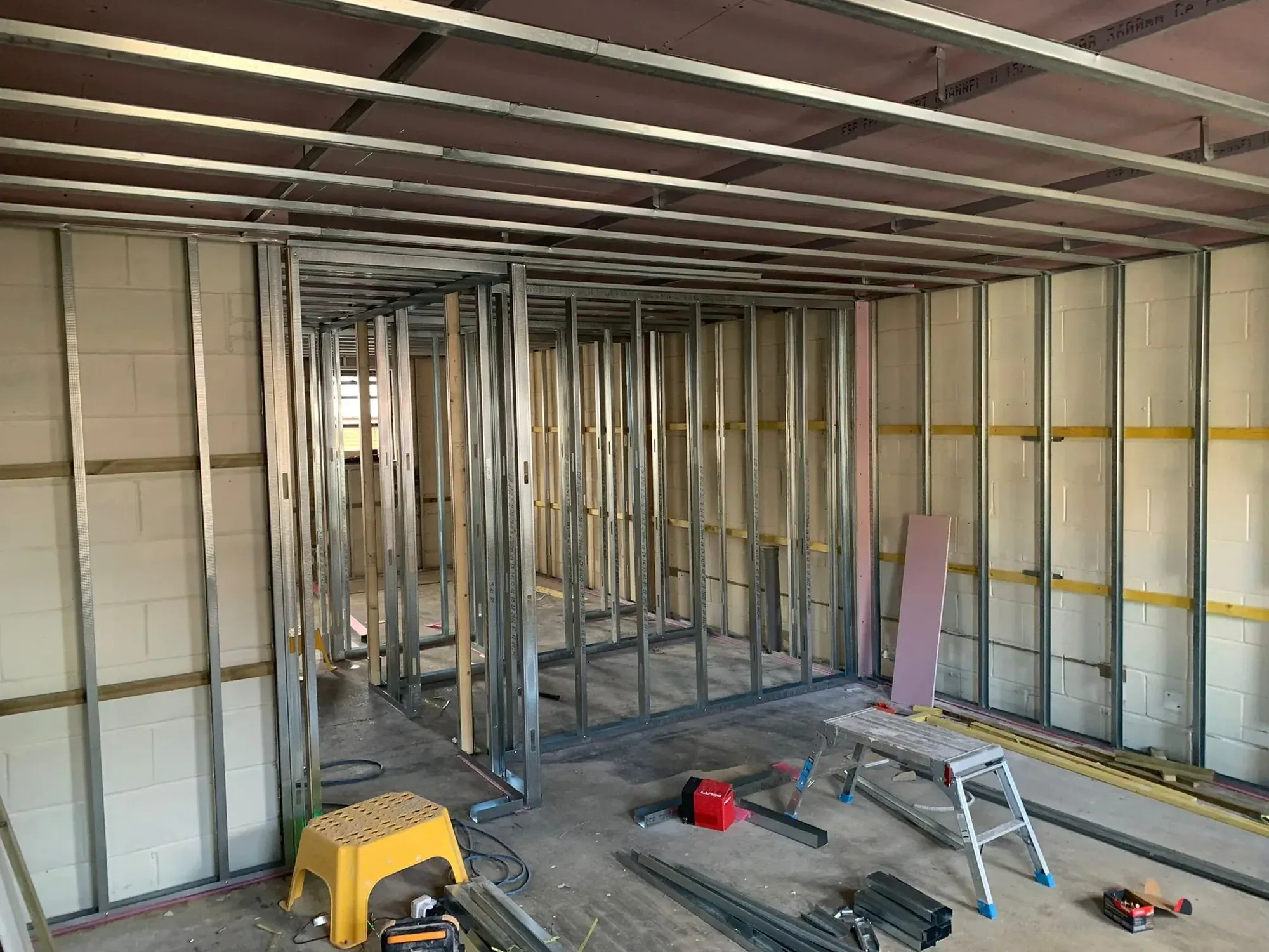 Metal stud partition framing set out through a ground floor, blockwork walls behind, tools and stepladders on the floor